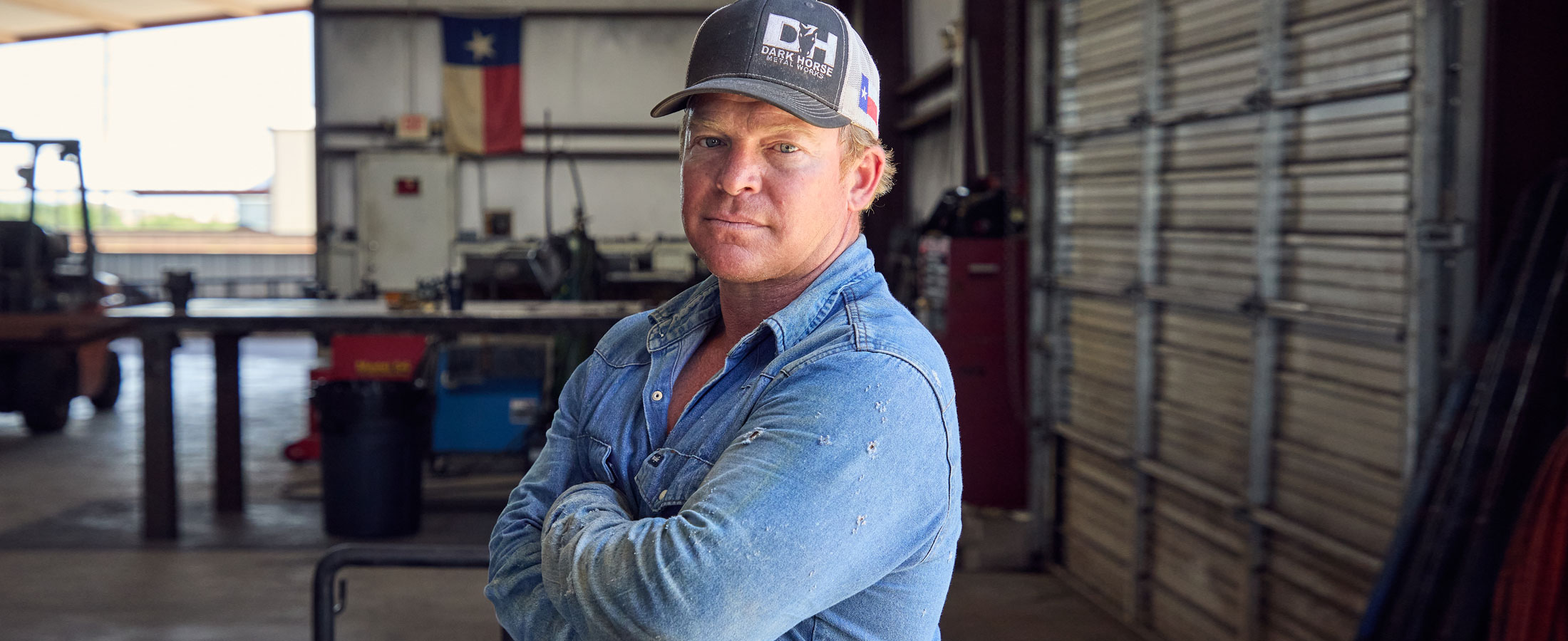 Un hombre, Wes Stevenson, con una camisa vaquera desgastada y una gorra de béisbol, está de pie con los brazos cruzados en un taller de metal, mirando con confianza a la cámara; la bandera de Texas cuelga de la pared detrás de él.
